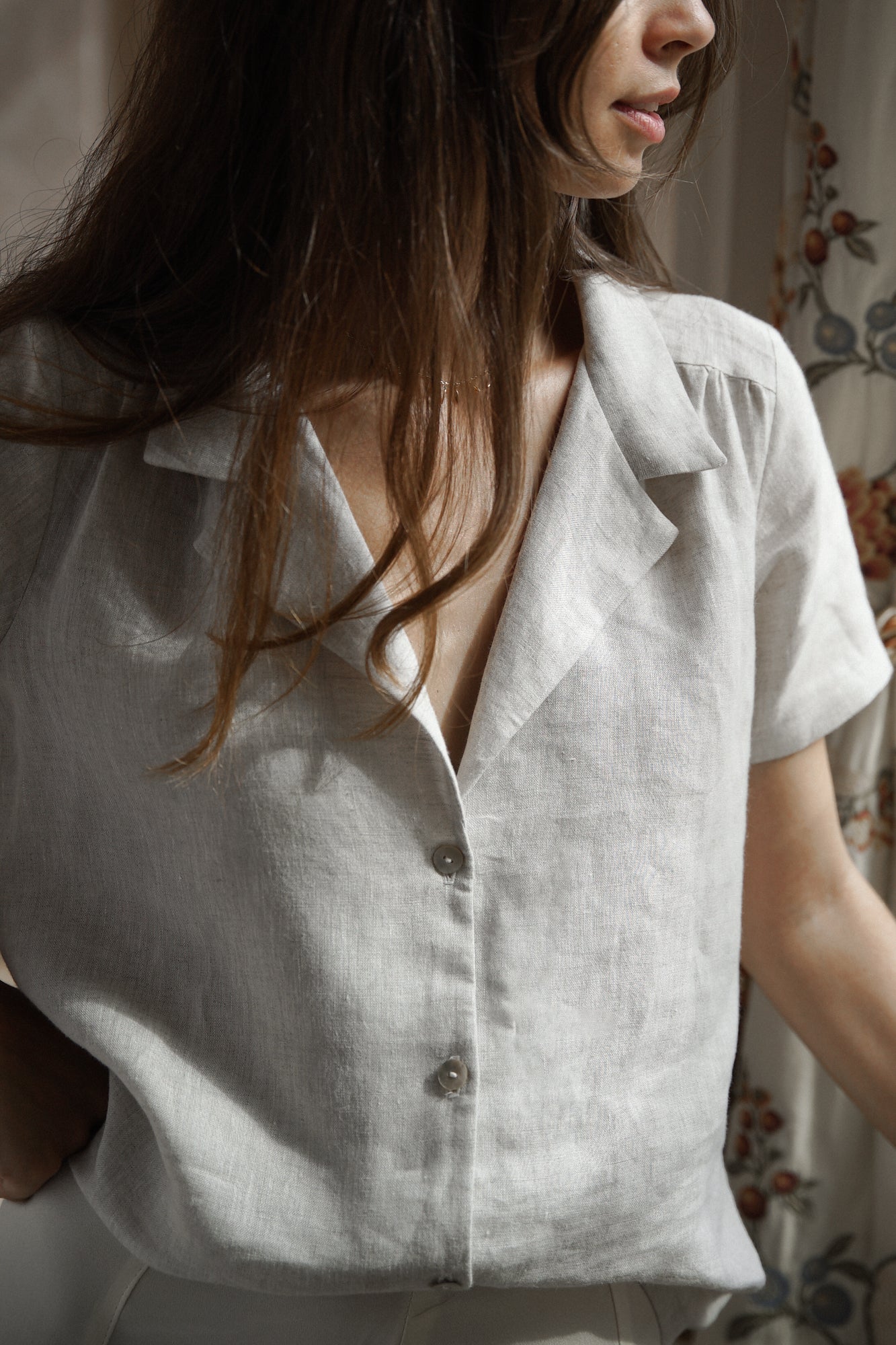 Femme portant une chemise en lin gris clair, avec un col tailleur minimaliste, des manches courtes ainsi que des boutons en nacre pour refermer sur le devant.
