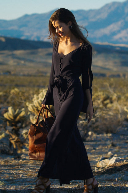 Femme portant une robe longue bleu, fluide et légère, au milieu d'un désert aride. Elle tient un sac de voyage à la main et regarde par terre. Les manches de sa robe sont longues et ajourées, on aperçoit des détails de peau sur la partie supérieur de la manche. Sa robe est cintrée par une ceinture en tissu bleu à la taille.