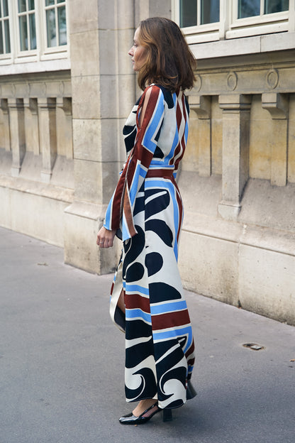 Femme dans une rue parisienne portant une longue robe maxi en polyester, avec des motifs rétro type psychédélique de couleur noir, marron, bleu et blanche. Les manches de la robe sont maxi et ajourées, ce qui fait qu'on observe des bouts de peau, et la robe est cintrée à la taille par une ceinture en tissu également.