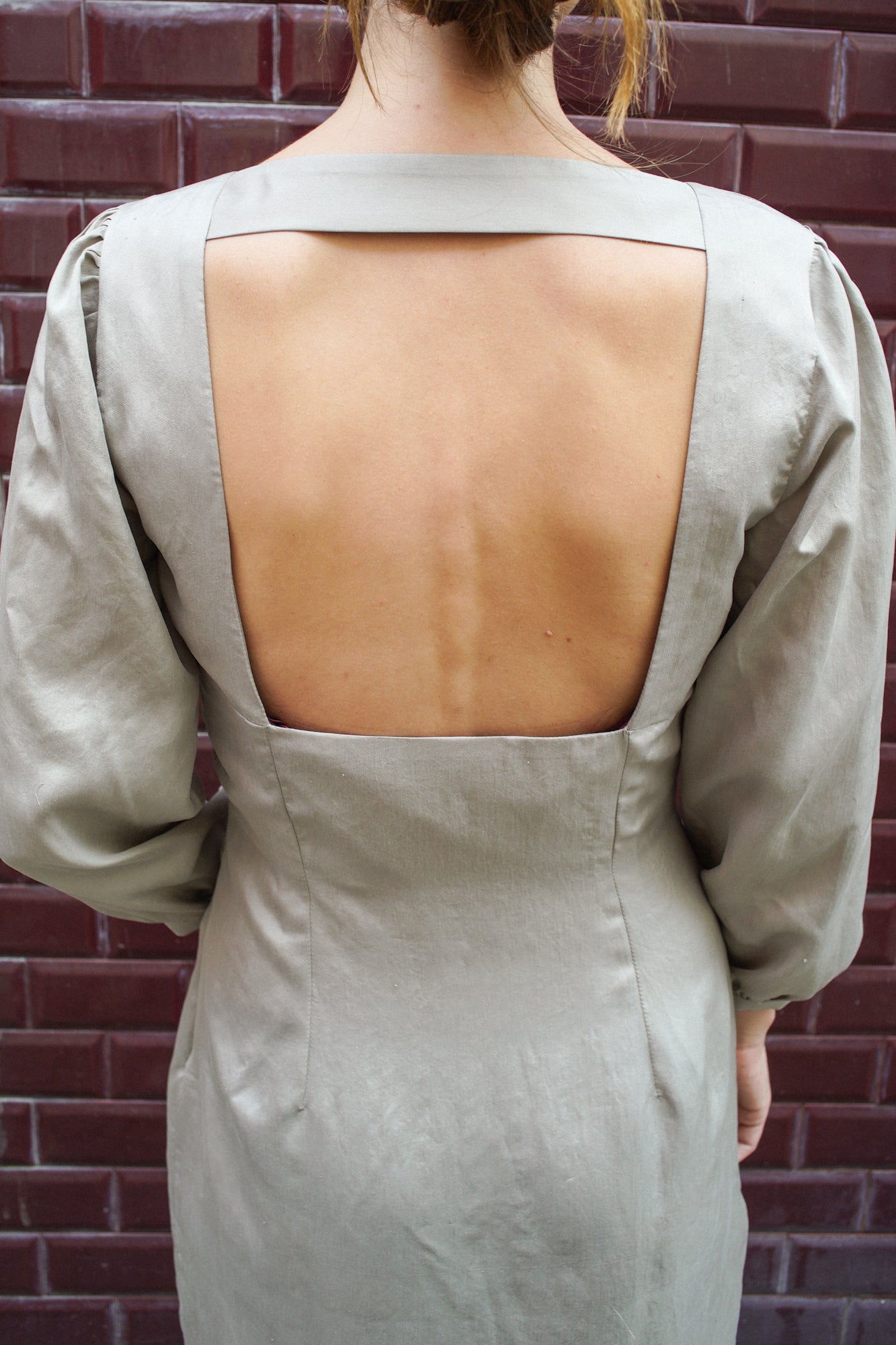 Zoom sur le dos nu d'une robe de couleur gris clair. Le dos nu est carré, très structuré par la présence d'une bande de tissu en haut du dos. On aperçoit également les manches de la robe qui sont longues, 3/4 et bouffantes.
