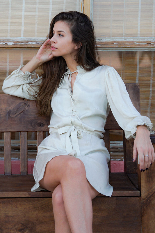 Jeune femme assise sur un banc, et portant une robe courte blanche avec des détails froufrous au niveau de l'encolure et des emmanchures. La robe est cintrée par une ceinture en tissu blanc.