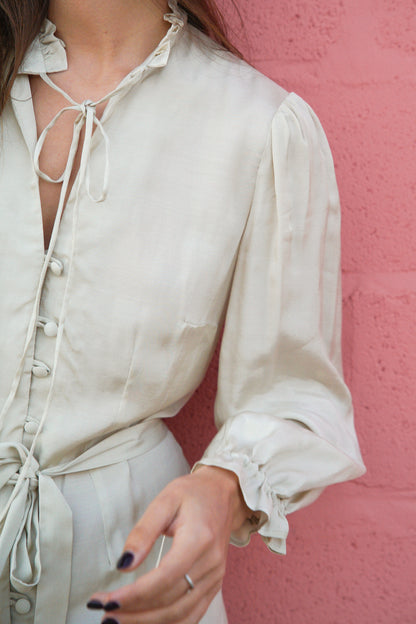 La photo est zoomée sur une robe courte blanche avec des détails froufrous au niveau de l'encolure et des emmanchures. La robe est cintrée par une ceinture en tissu blanc et se ferme par des boutons recouverts de tissu.