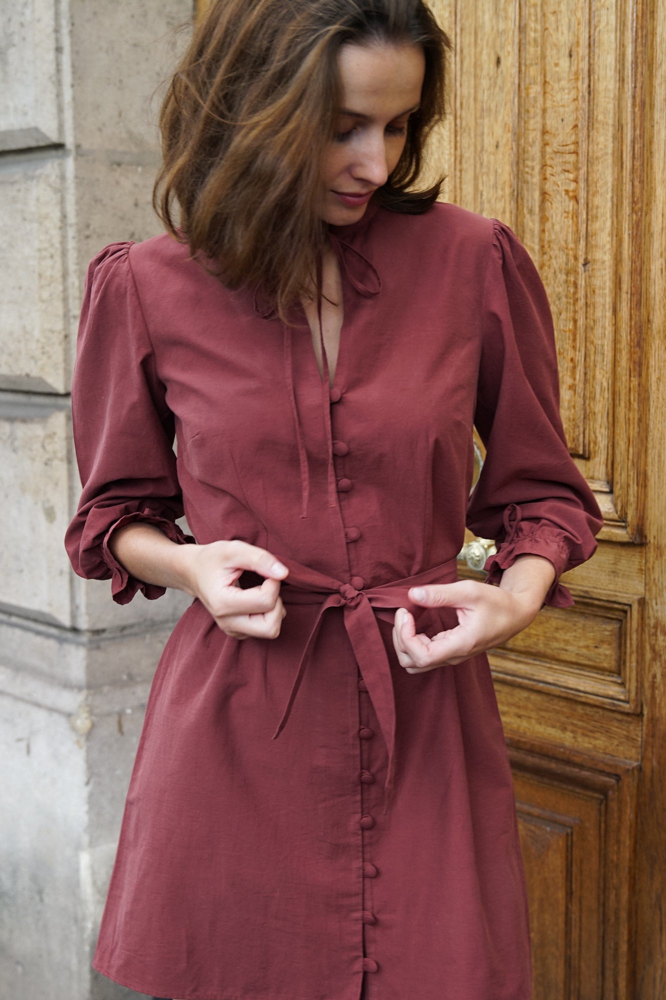 Femme portant une robe courte de couleur rouge/marron, devant une porte d'immeuble parisien. La robe est cintrée à la taille, avec une ceinture en tissu et des boutons recouverts sur le devant. Elle a des détails froufrous sur le col et sur ses manches bouffantes, ainsi que des fermetures à brides. C'est un modèle de la marque éthique et éco-responsable Pelmel Paris.