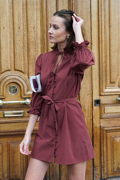 Femme portant une robe courte de couleur rouge/marron, devant une porte d'immeuble parisien. La robe est cintrée à la taille, avec une ceinture en tissu et des boutons recouverts sur le devant. Elle a des détails froufrous sur le col et sur ses manches bouffantes, ainsi que des fermetures à brides. C'est un modèle de la marque éthique et éco-responsable Pelmel Paris.
