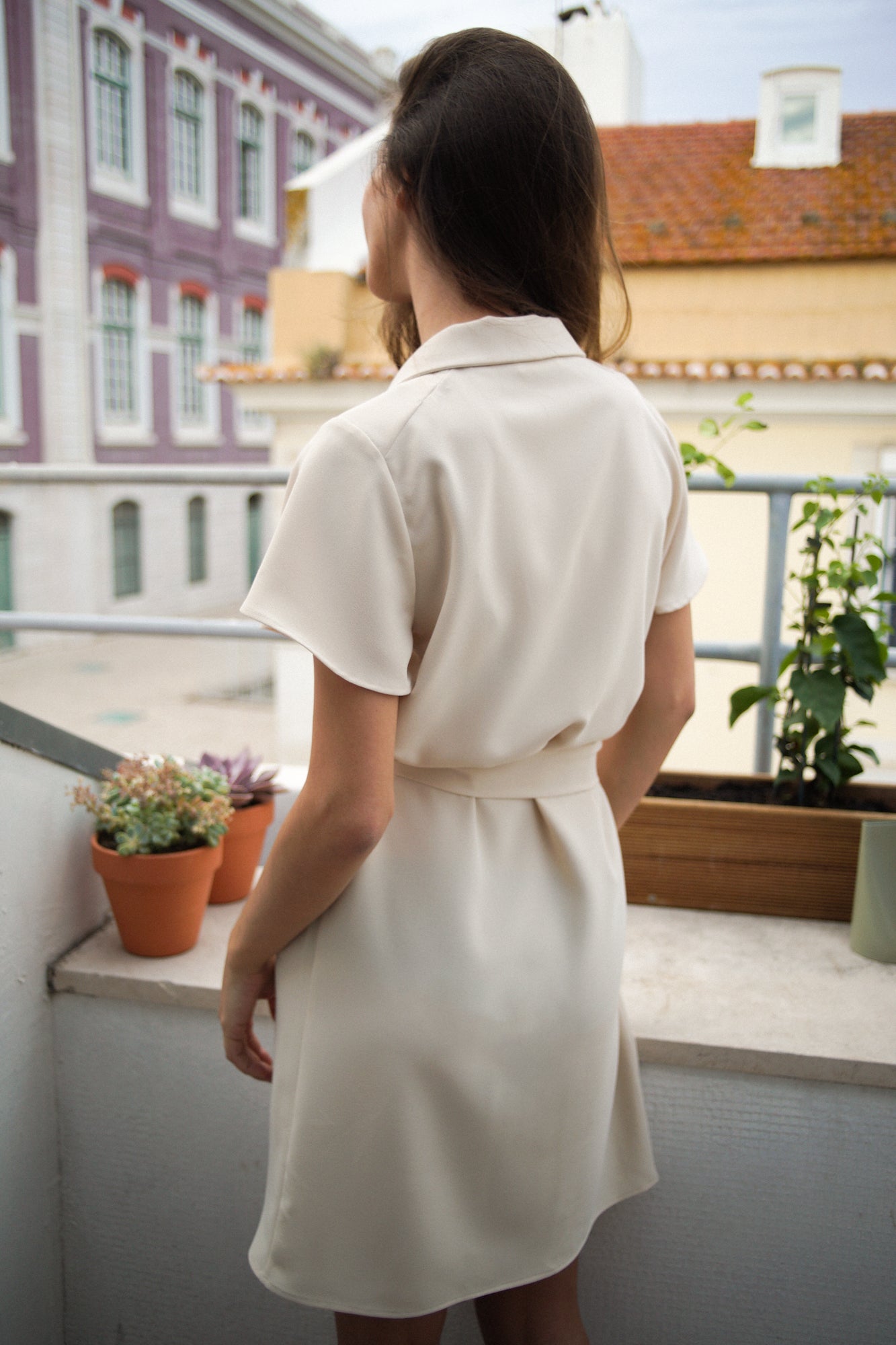 Femme de dos, sur un balcon d'appartement, portant une robe courte crème/blanc cassé. La robe possède des manches courtes simples et fluides, et un col. Elle est cintrée à la taille grâce à une ceinture en tissu.