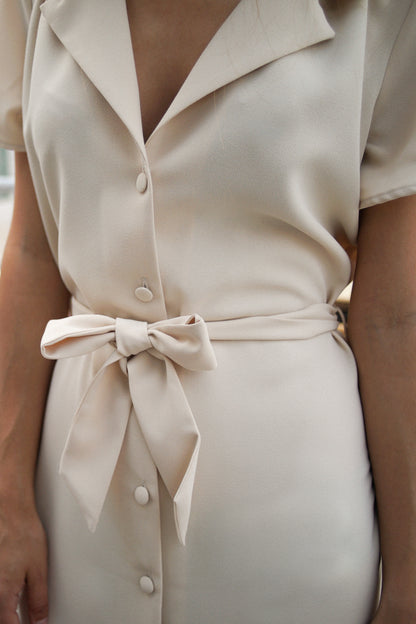 Zoom sur une robe blanc cassé/crème. La robe a des manches courtes et fluides, et se cintre à la taille par une ceinture en tissu et des passants. Elle se ferme par des boutons recouverts de tissu. Il s'agit d'un polyester tombant et fluide.