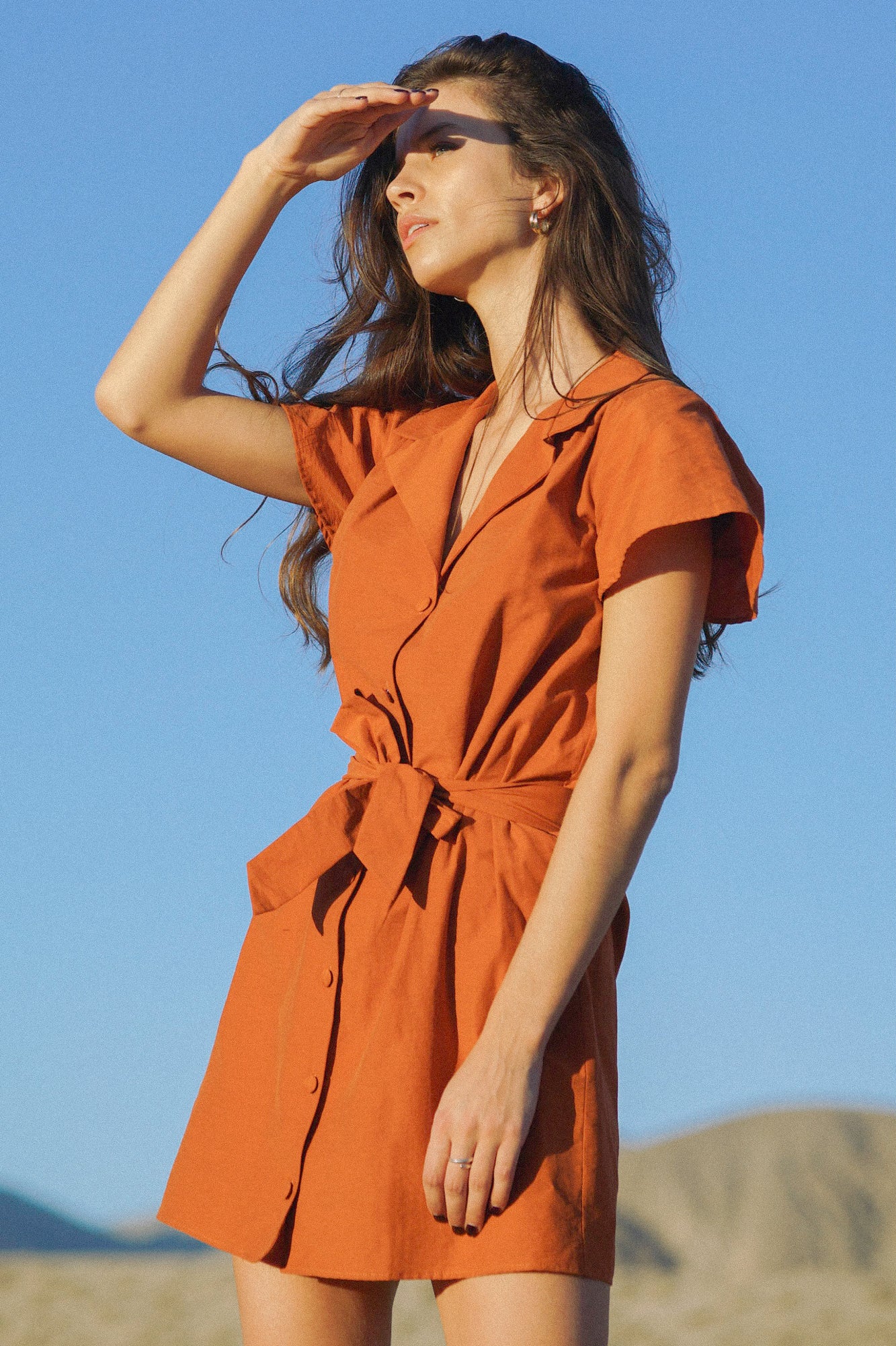 Robe orange/irisé portée par une femme au milieu d'un désert aride et avec le ciel bleu en arrière plan. La robe est courte, de couleur orange, cintrée par une ceinture en tissu à la taille, et comportant des boutons recouverts de tissus sur le devant pour la refermer. Les manches sont courts et simples, et le col de la robe est un col tailleur.