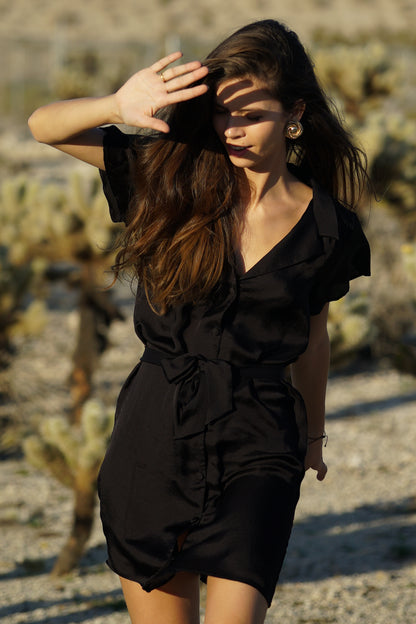 Femme debout au milieu du désert portant une robe noire courte. Elle se protège du soleil avec sa main droite. Sa robe courte est de couleur noire, cintrée à la taille grâce à une ceinture en tissu noire. La robe a des manches courtes et fluides, un col tailleur ainsi que des boutons recouverts tissus pour la fermer sur le devant.