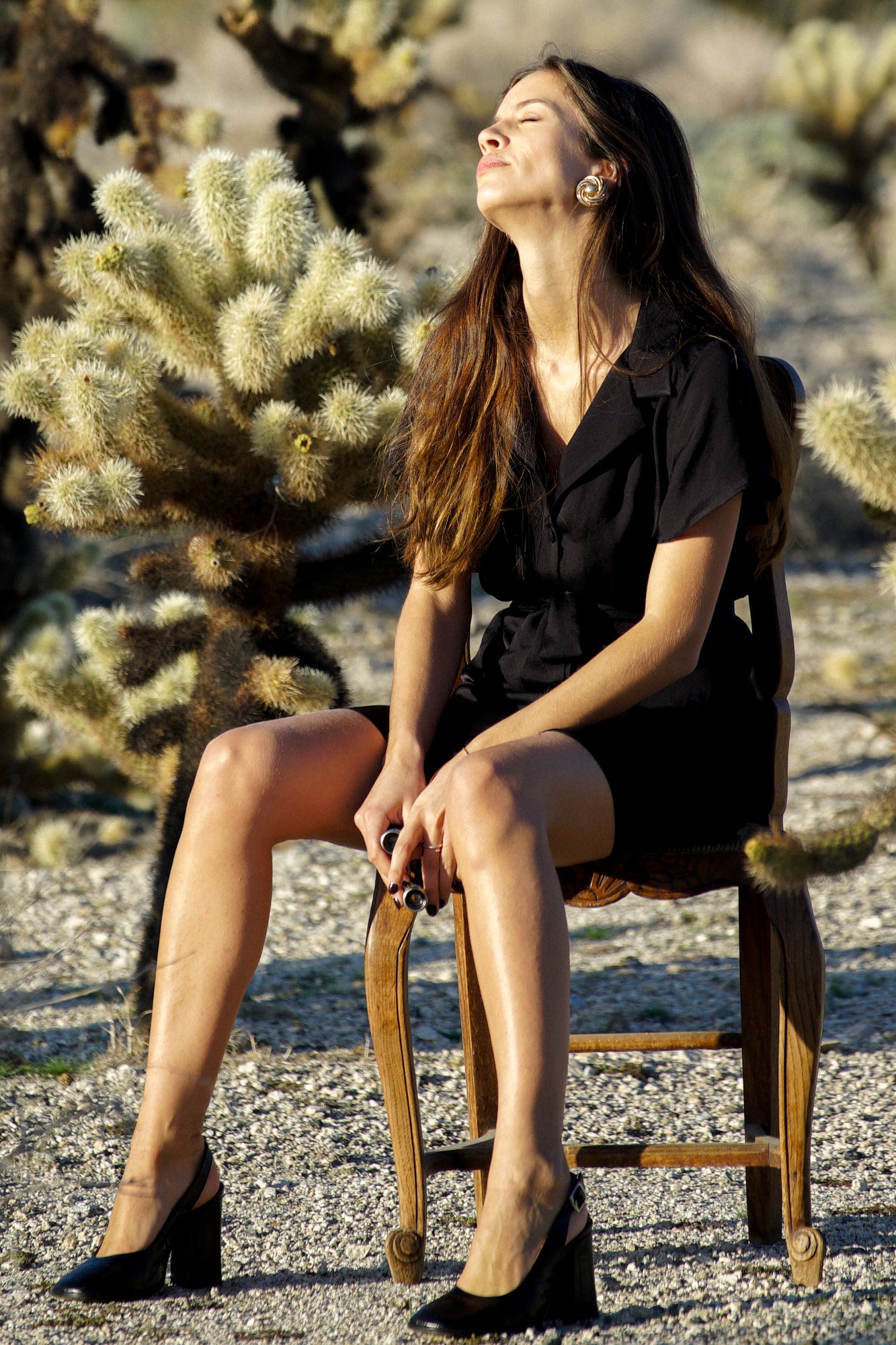 Femme sur une chaise et tournants sa tête pour bronzer au soleil. Elle porte une robe courte noire, avec des manches courtes et un col tailleur minimaliste. La robe se ferme par des boutons recouverts de tissu sur le devant, et elle est composée de polyester soyeux et fluide.