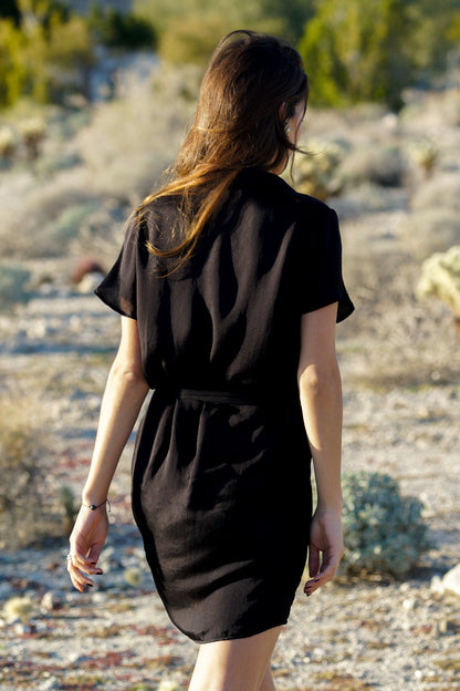Femme marchant de dos au milieu d'un désert aride. Elle porte une robe noire courte, avec des manches courtes fluides et légères, ainsi qu'une ceinture à la taille en tissu. La robe a l'air fluide et très agréable à porter.