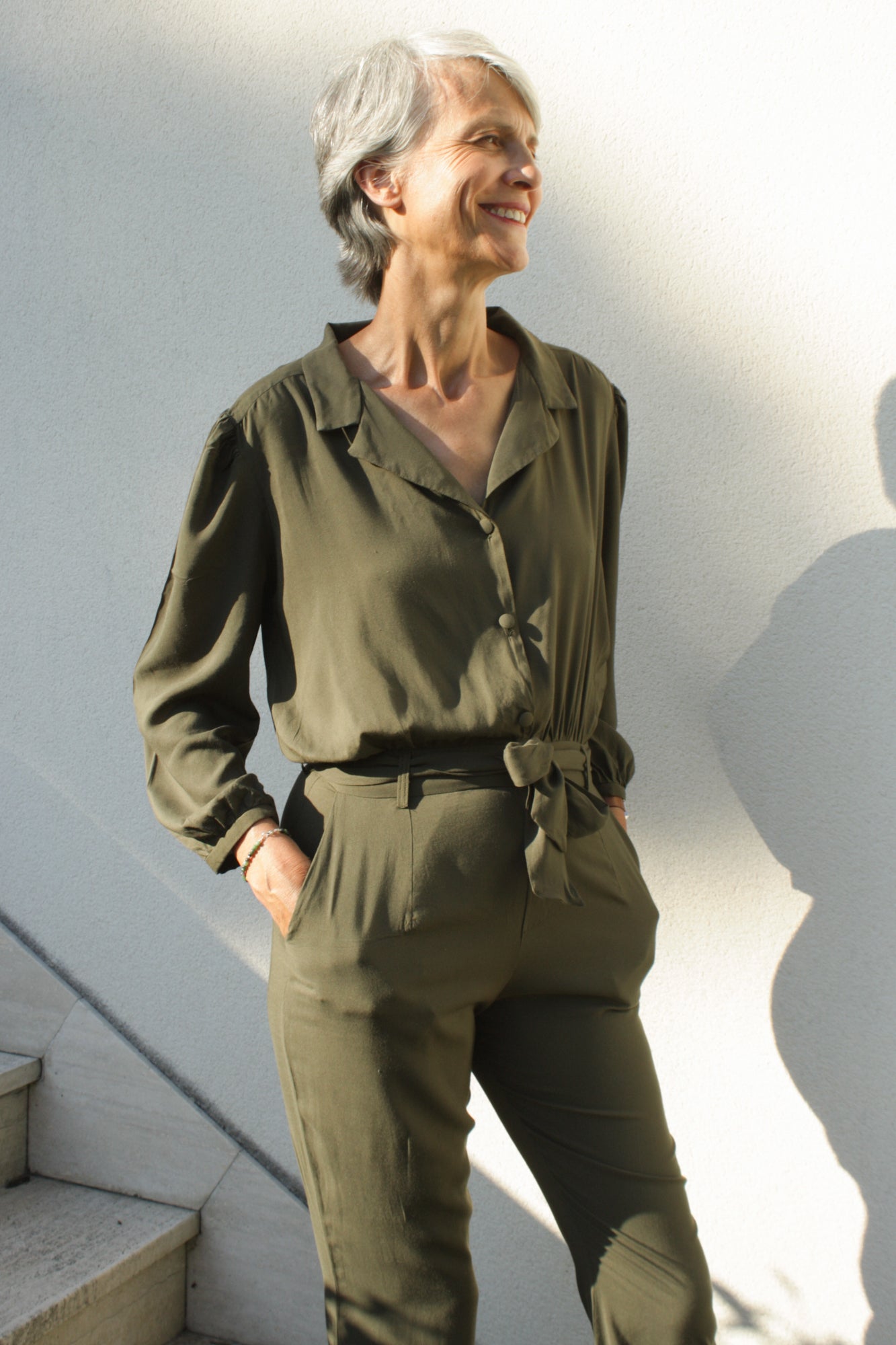 Femme mûre portant une combinaison vert kaki, avec une ceinture en tissu à la taille. La combinaison est en viscose, sous la forme d'un tissu léger et fluide. Elle comporte un col raille élégant ainsi que des manches longues 3/4.