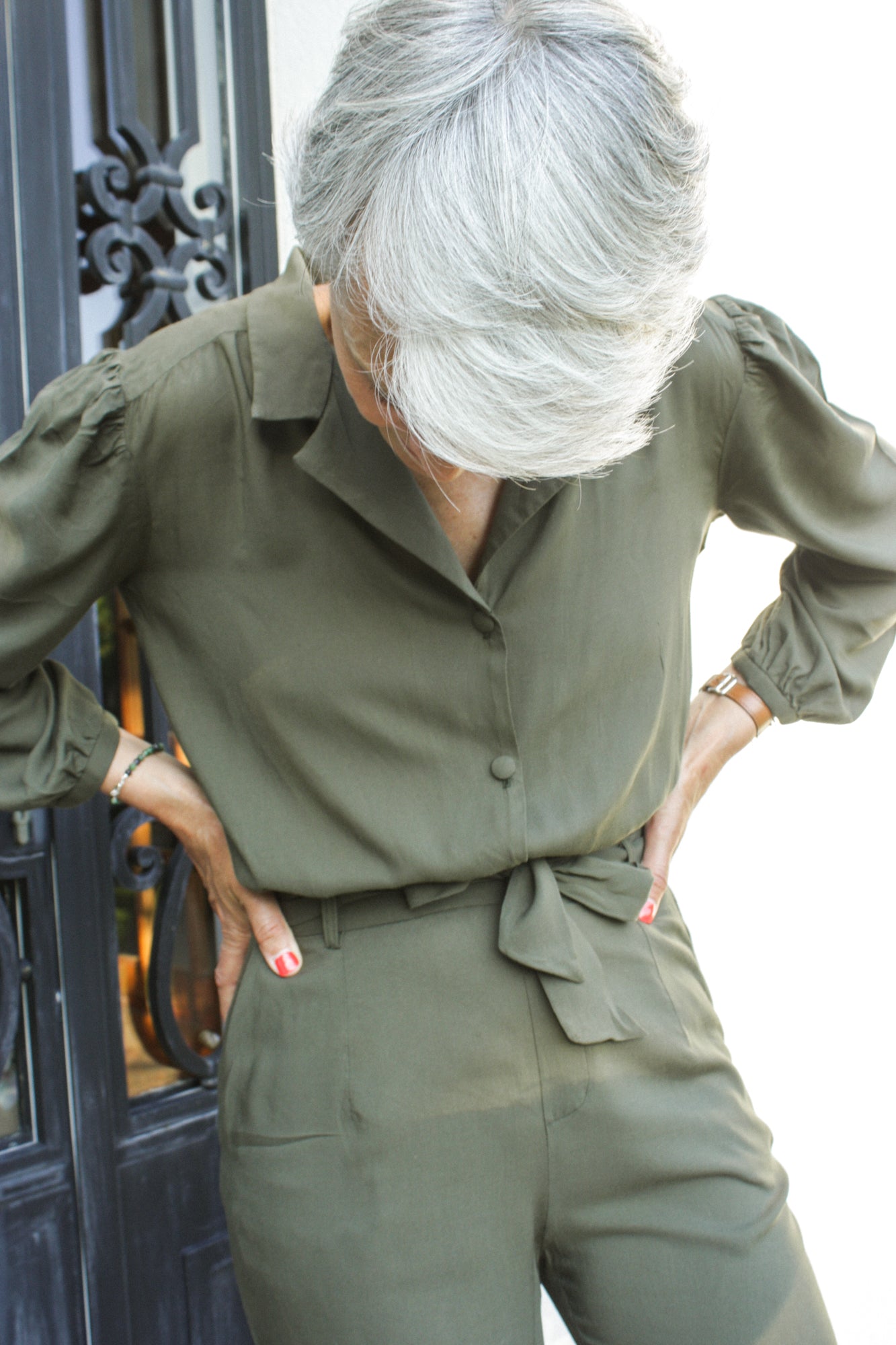 Femme mûre portant une combinaison vert kaki, avec une ceinture en tissu à la taille. La combinaison est en viscose, sous la forme d'un tissu léger et fluide. Elle comporte un col raille élégant ainsi que des manches longues 3/4.