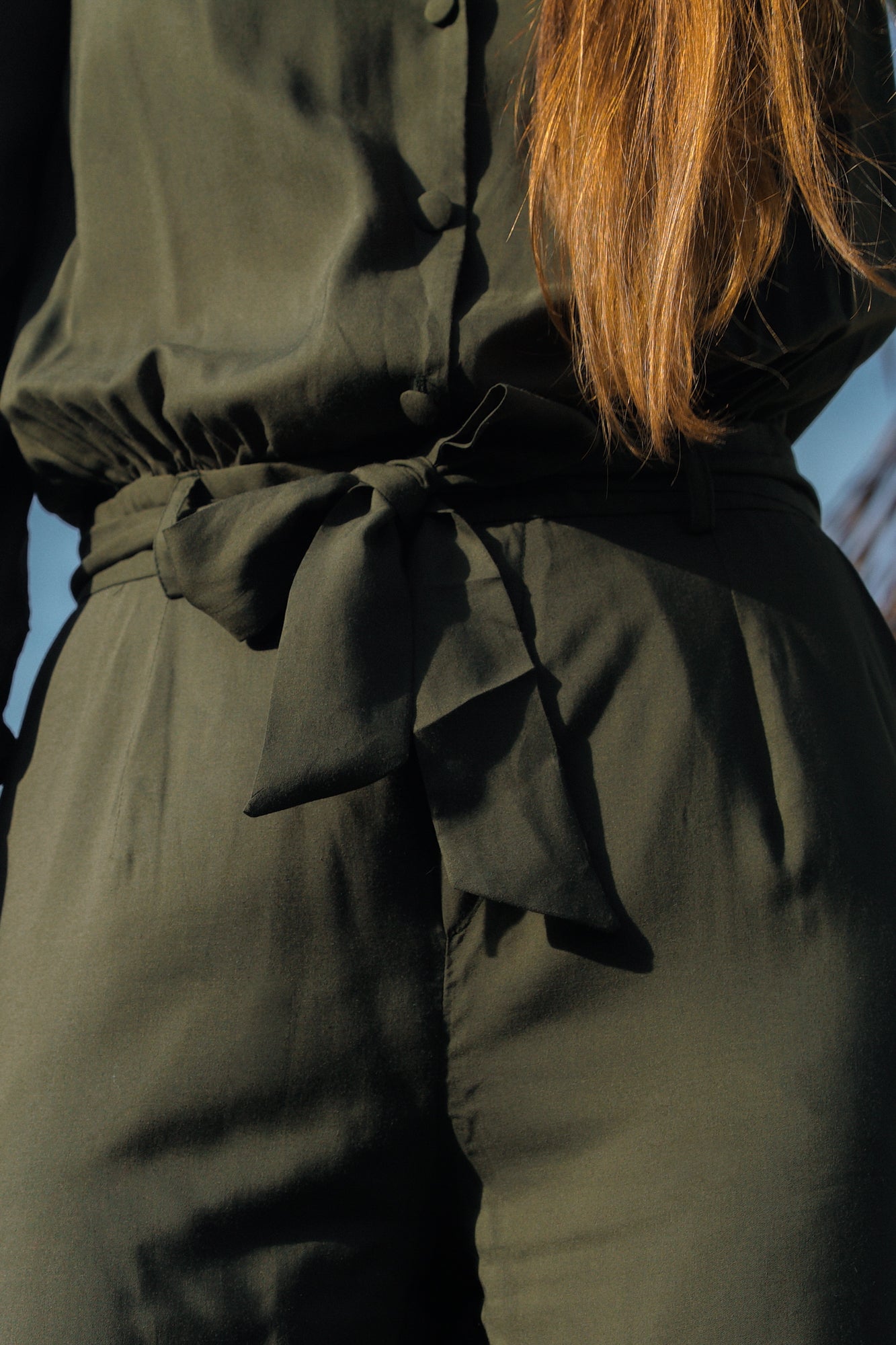 Zoom sur la ceinture en tissu d'une combinaison longue vert kaki. La femme qui porte la combinaison a les cheveux longs détachés. La combinaison comprend des boutons recouverts de tissus sur le devant, et on peut voir que le tissu a l'air très agréable au touché et soyeux.